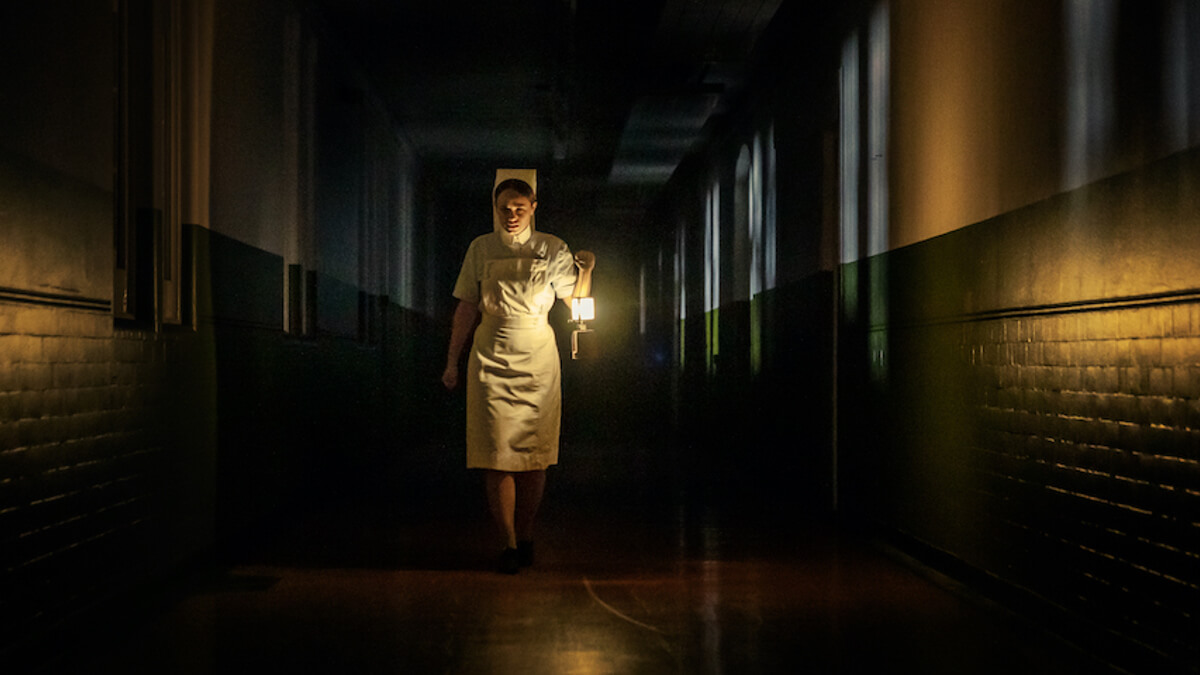 Enfermera con un faro de luz caminando en un pasillo oscuro en la película La oscuridad 
